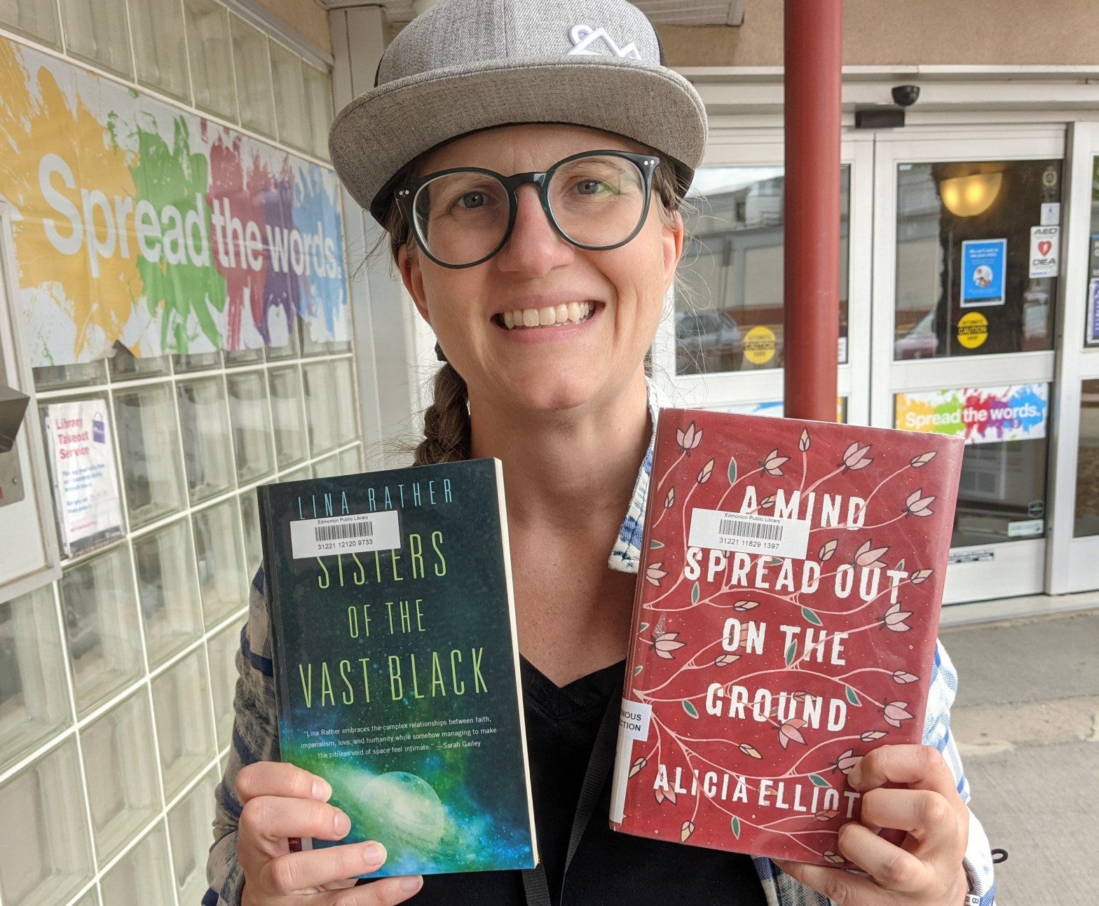a woman wearing a grey ball cap and blue round glasses is holding up two books to the camera. the book on the left has an image of outerspace on it. the book on the right is a rich red colour with dainty drawings of leaves and flowers on it with a white title filling most of the cover.