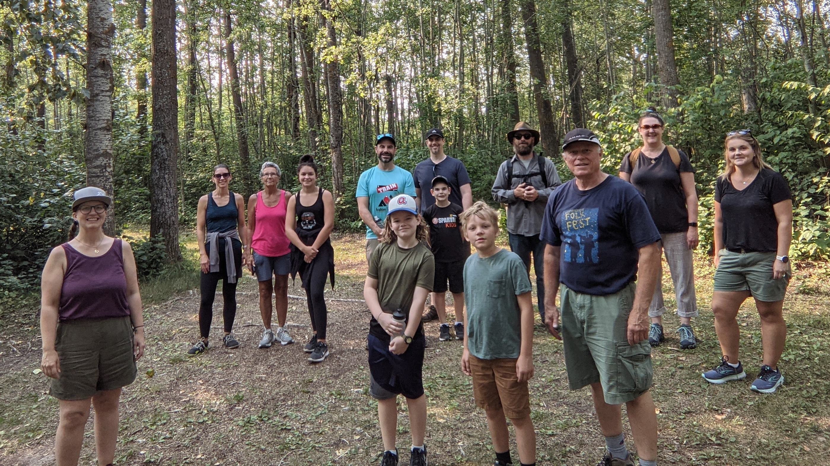 Group of 13 people dressed in summer outdoor clothing in a clearing surrounded by a grove of deciduous trees