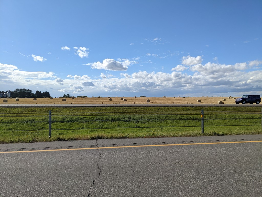 A very scenic image. Bottom third is an asphalt highway, middle is green grass and a beige field with hay bales dotted throughout. And the top third is a bright blue sky with white puffy clouds that go no for miles.