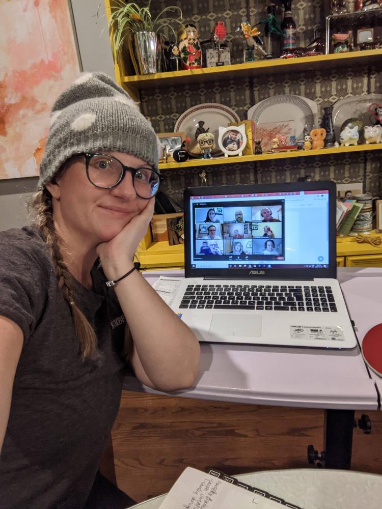 Selfie of a white woman in a grey toque sitting at a purple desk with white laptop on it. The screen shows a virtual meeting. There is a curio cabinet behind her.