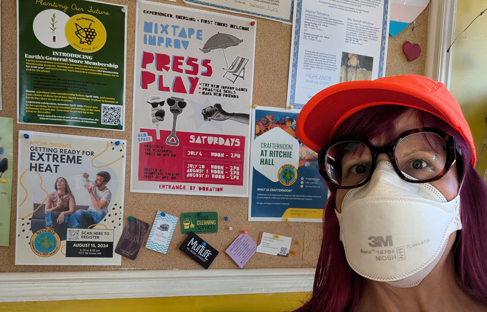 Lisa in her orange ball cap and mask stands in front of a bulletin board covered with colourful posters